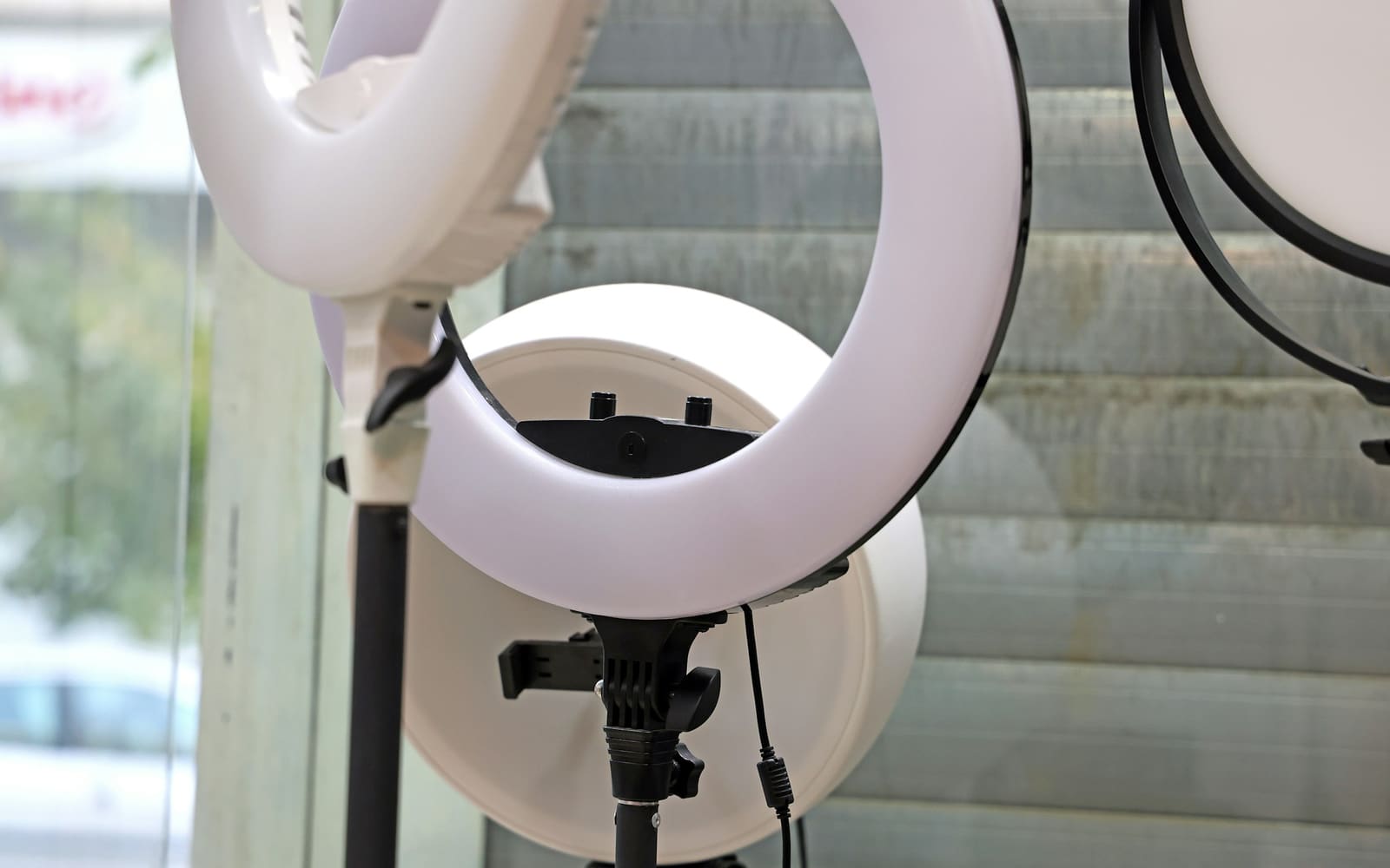 Three white ring lights on black stands, the middle one in front of a round white panel, beside a window with gray slatted blinds