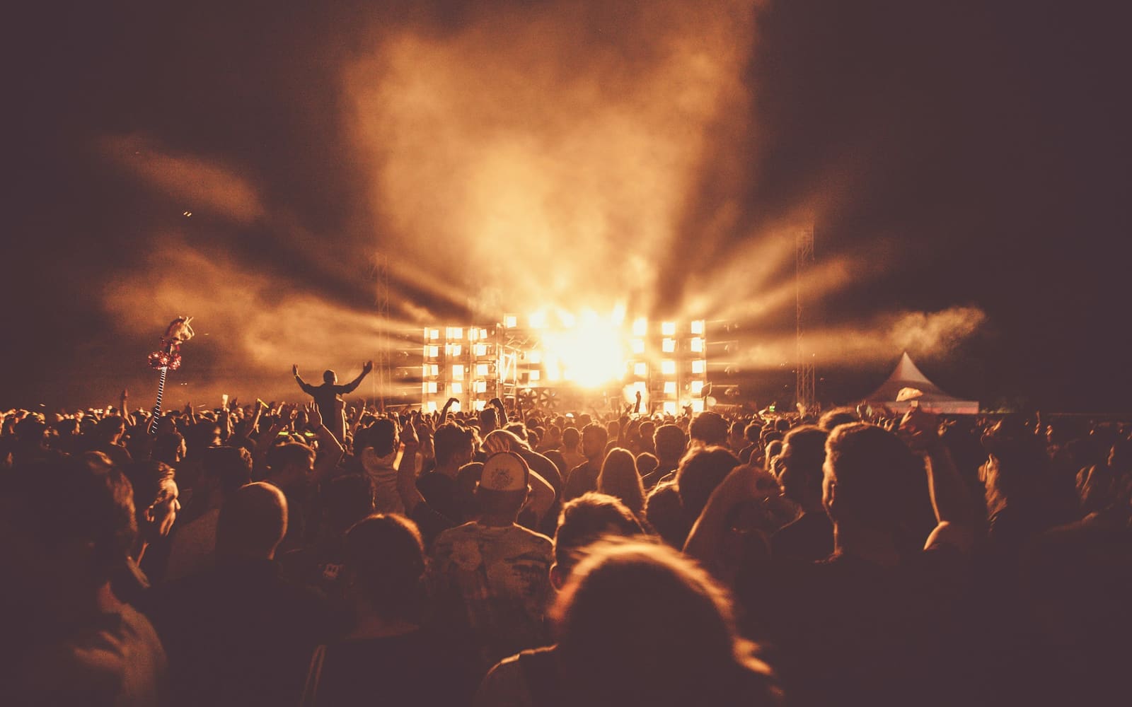 A large crowd seen from behind faces a festival stage at night, with beams of warm orange light and smoke rising into a dark sky