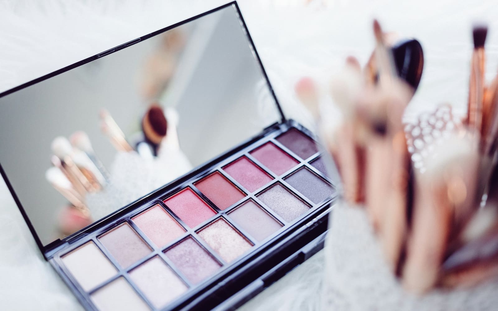 An open eyeshadow palette of pink, mauve and nude shades with its mirror raised, beside a blurred cup of makeup brushes on white fur