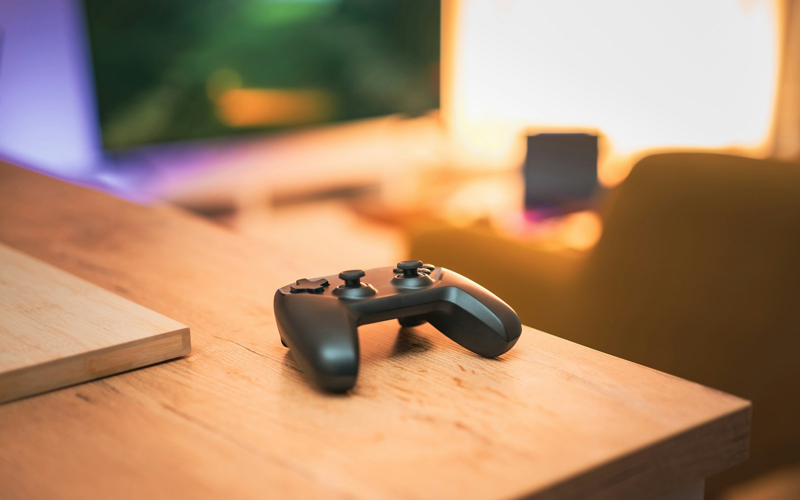 A black game controller resting on the edge of a light wooden desk, with a blurred monitor and warm lamp light behind it
