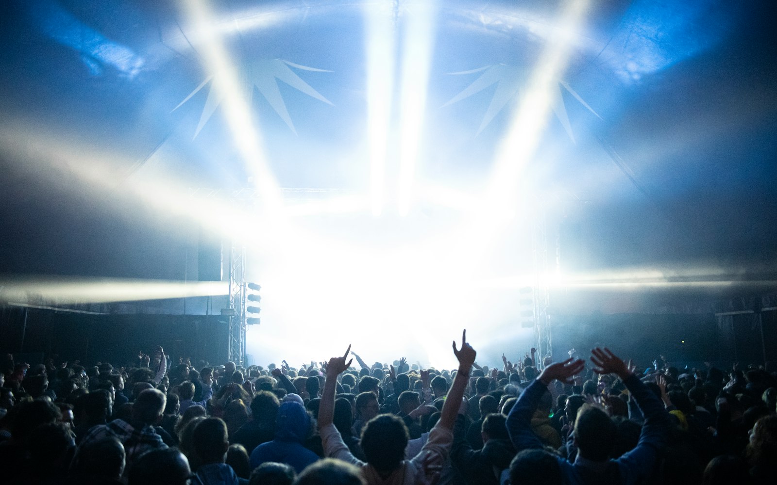 A crowd seen from behind with raised hands, facing a stage flooded with bright white and blue light beams under a tent roof