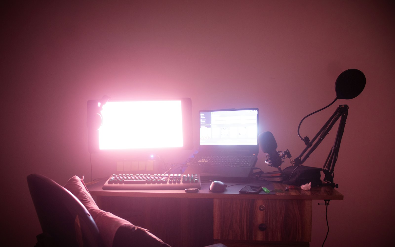 A small wooden desk in a dim, pink-lit room with a glowing monitor, a laptop, a keyboard and a microphone on a boom arm