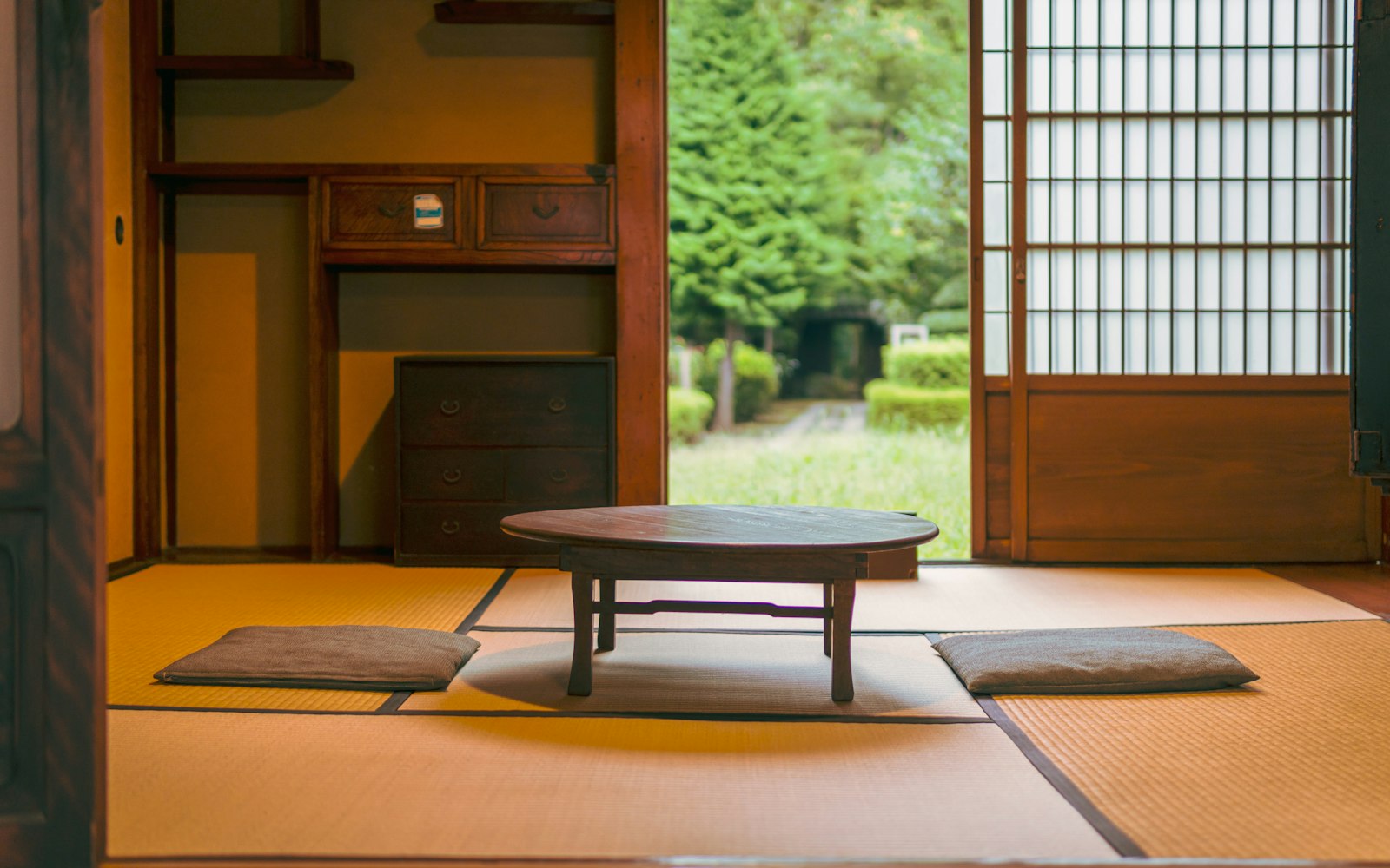A traditional Japanese room with tatami mats, a low round wooden table and two floor cushions, its sliding door open to a garden