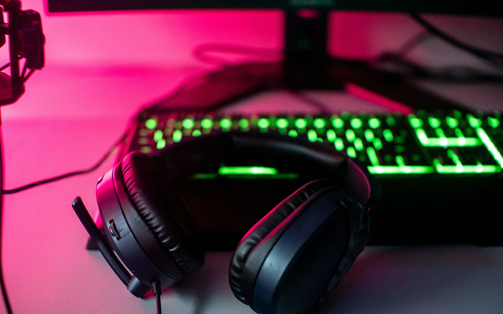 A black over-ear gaming headset with a boom microphone resting in front of a green-backlit keyboard on a desk lit with pink light