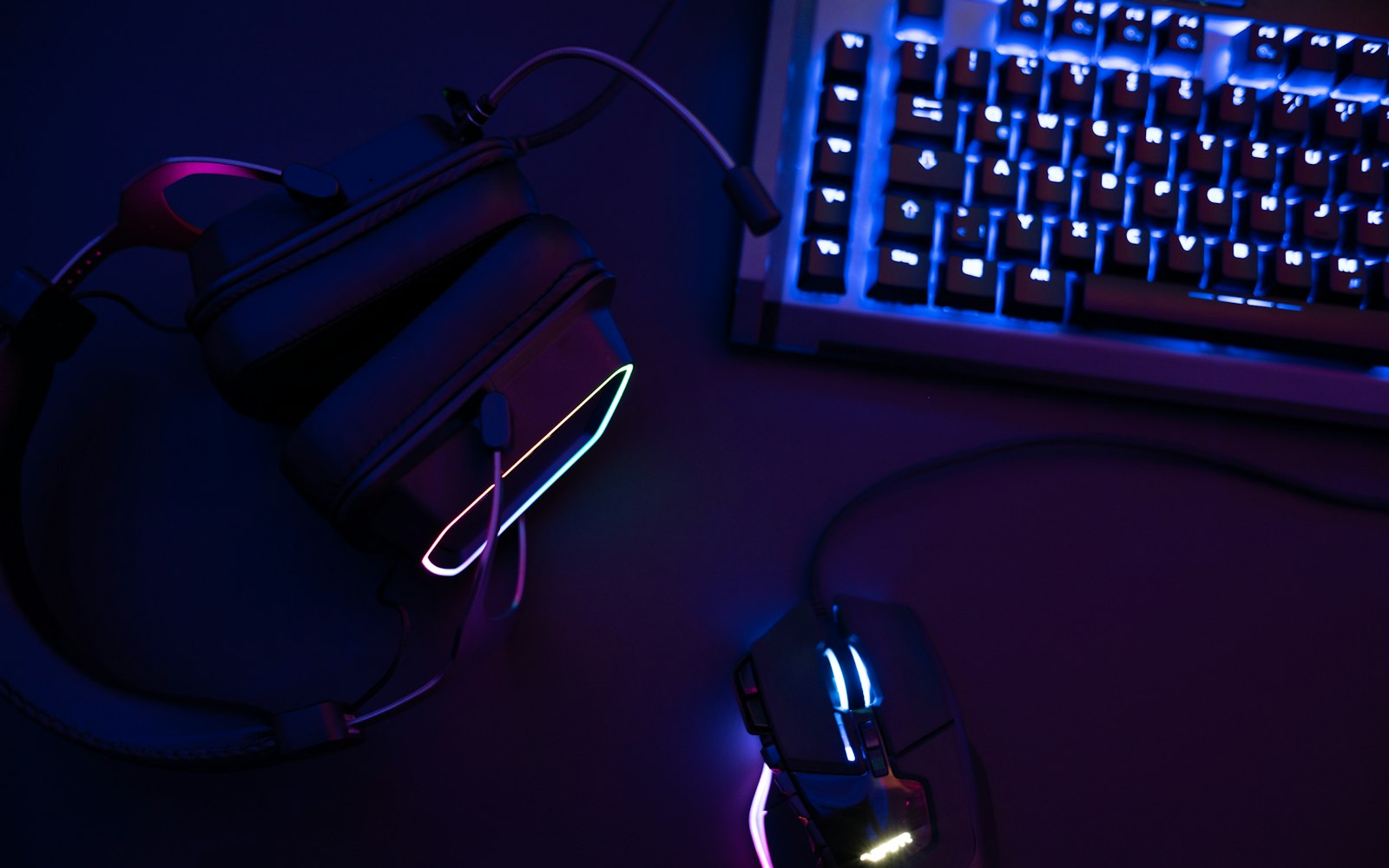 Overhead view of a dark desk with a blue-backlit keyboard, a glowing gaming mouse and a headset with a pink and cyan light strip