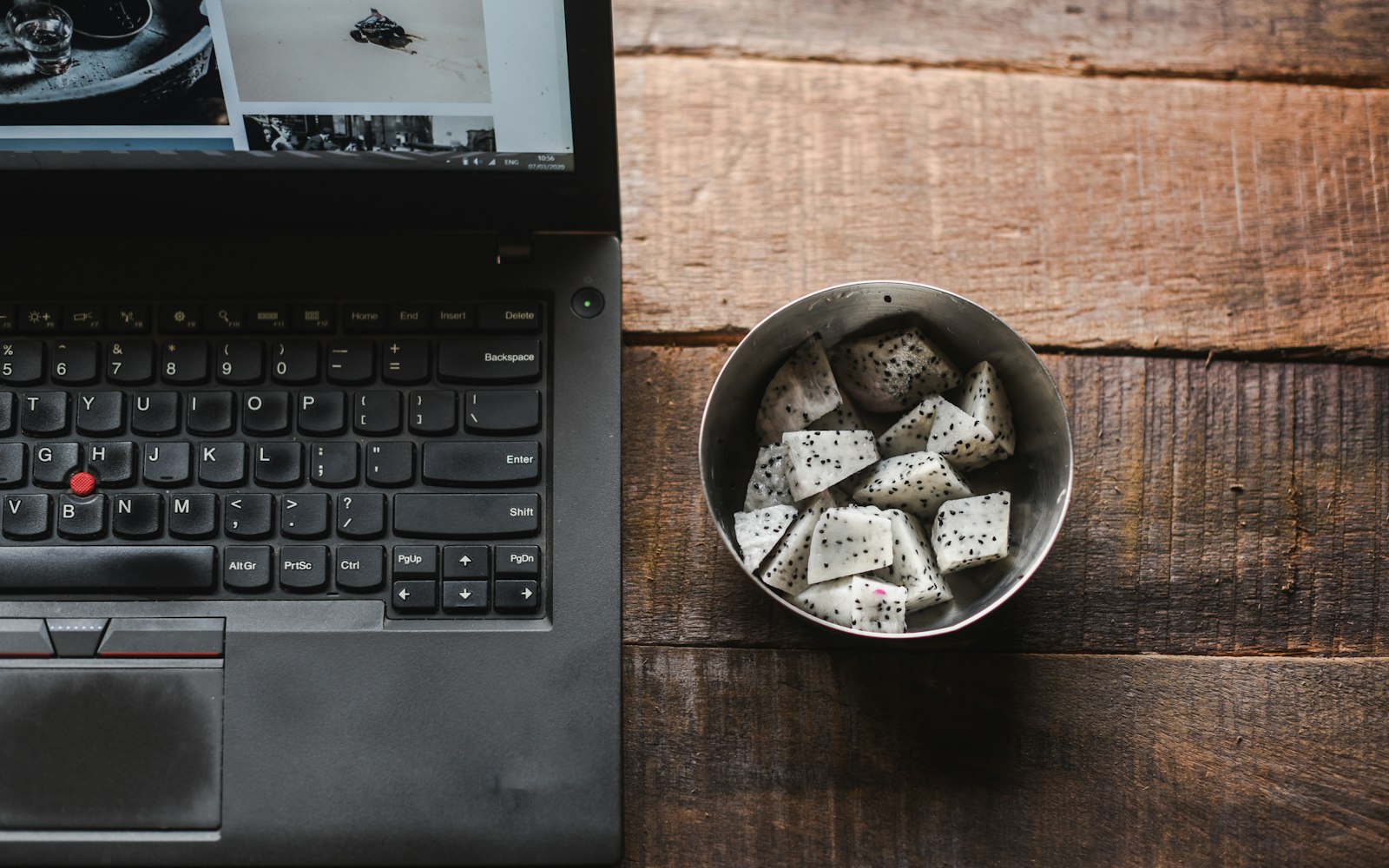 Top-down view of a black laptop keyboard on a rustic wooden table beside a metal bowl of cut white dragon fruit