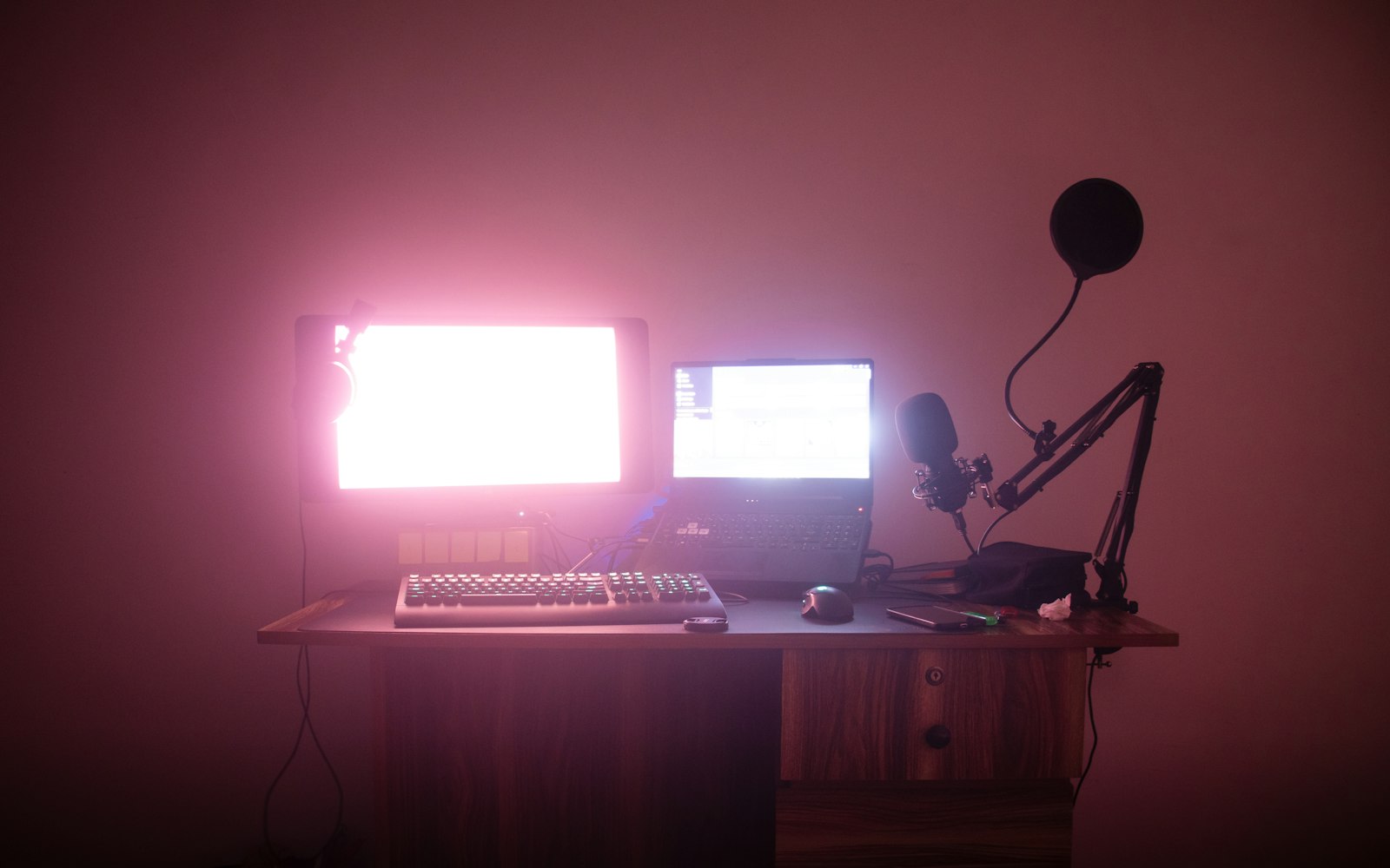 A pink-lit desk with a glowing monitor, an open laptop, a keyboard, a mouse and a boom-arm microphone with a pop filter