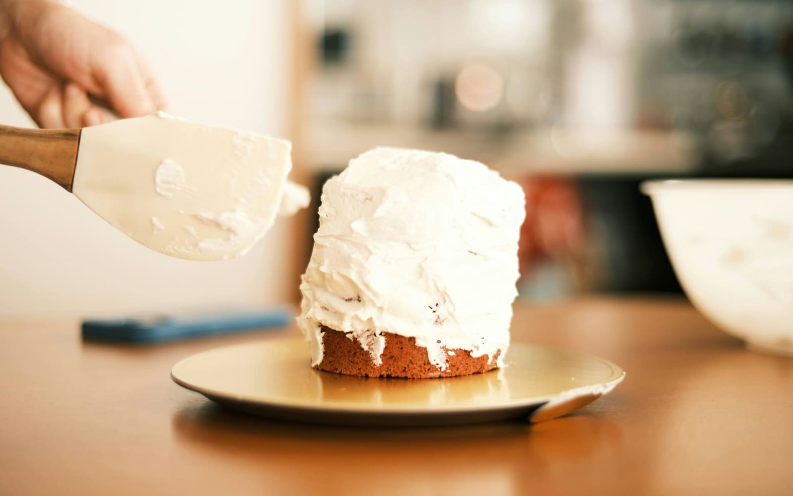 A hand holds a wooden-handled spatula beside a small cake thickly covered in white frosting on a gold board, with a mixing bowl nearby