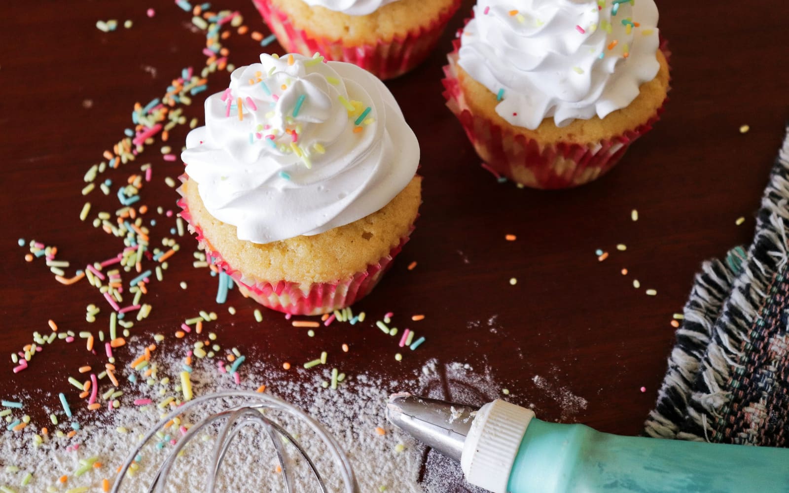 Three cupcakes with swirls of white frosting and pastel sprinkles on a dark wooden table beside a whisk and a teal piping bag