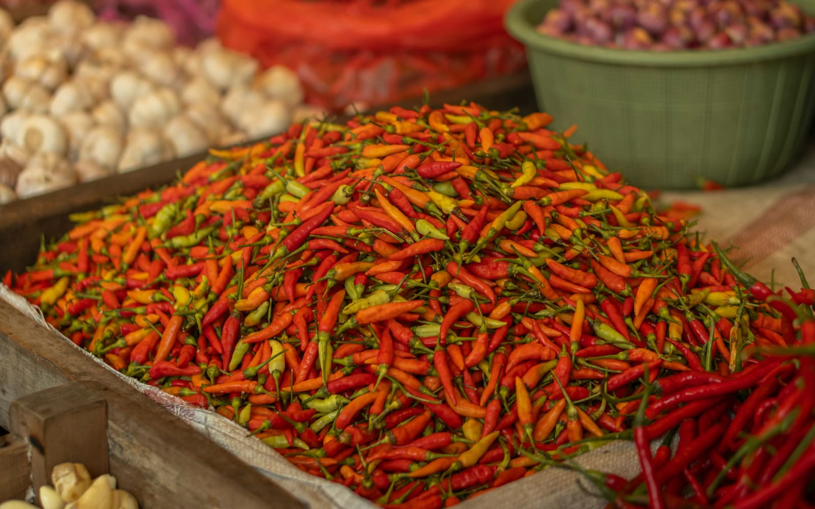 A heap of small red, orange and yellow chili peppers fills a wooden market crate, with garlic bulbs and a green basin of shallots behind