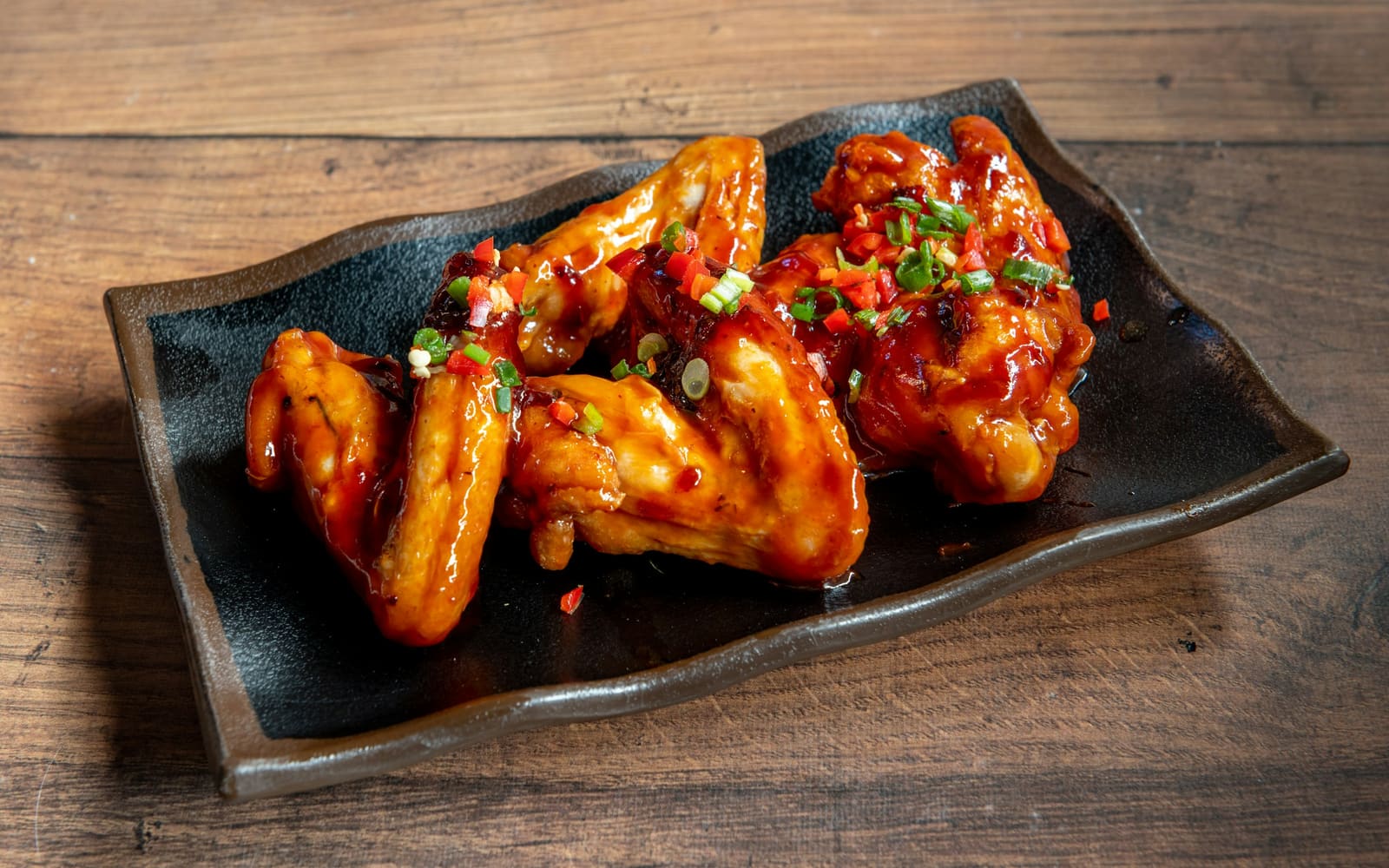Glossy chicken wings coated in a red-orange glaze and sprinkled with chopped chili and green onion sit on a dark plate on a wooden table
