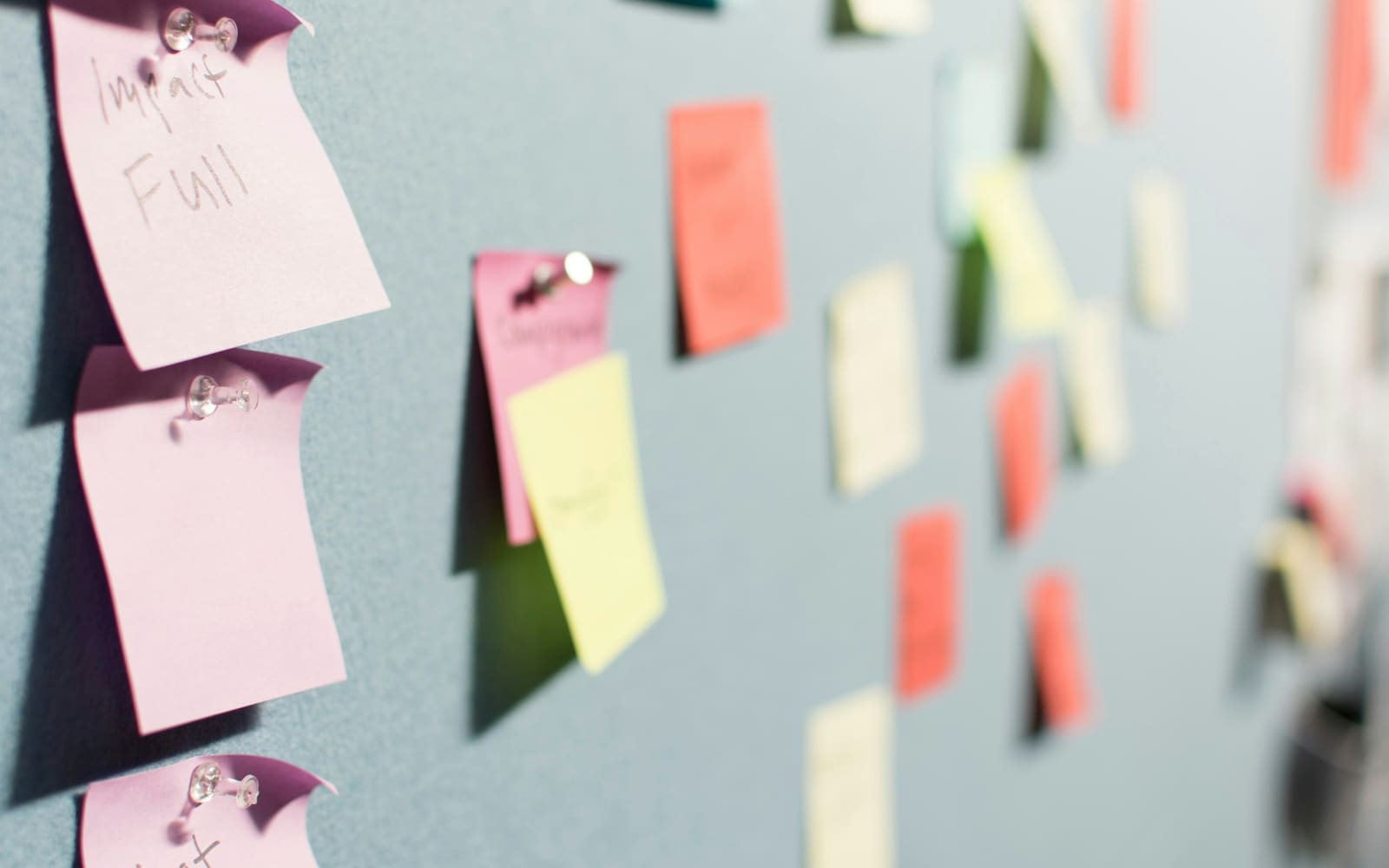Pink, yellow, orange and cream sticky notes pinned with clear pushpins to a gray-blue board, the nearest note handwritten, the rest blurred