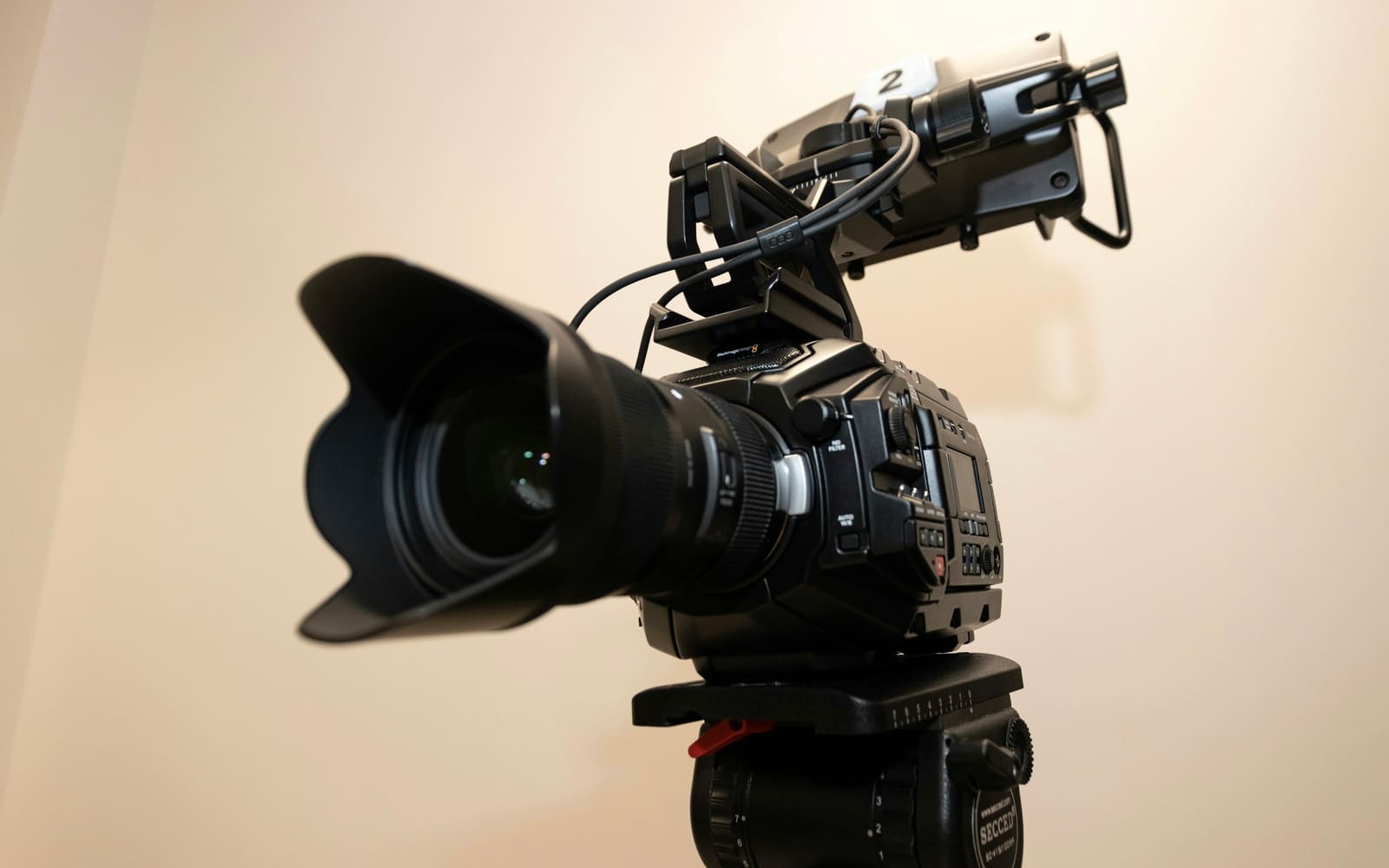A black video camera with a long lens hood on a tripod head, a small number 2 label on its top handle, against a cream wall