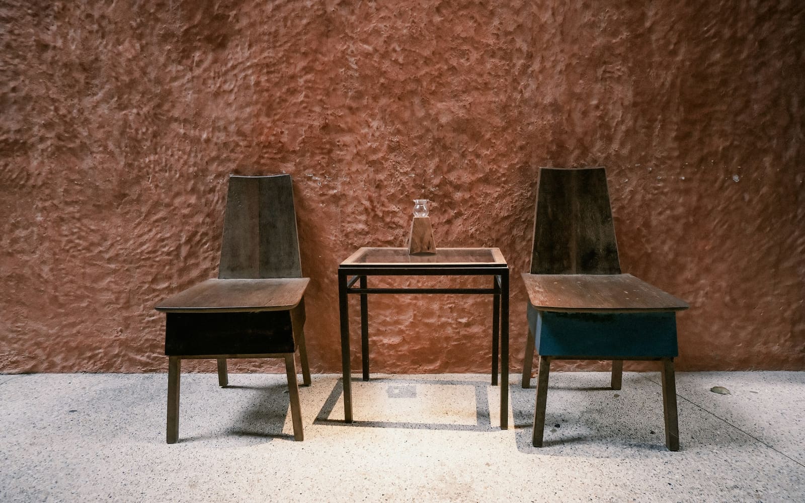 Two wooden chairs, one with a teal base, stand on either side of a small glass-topped table holding a carafe, against a rough terracotta wall