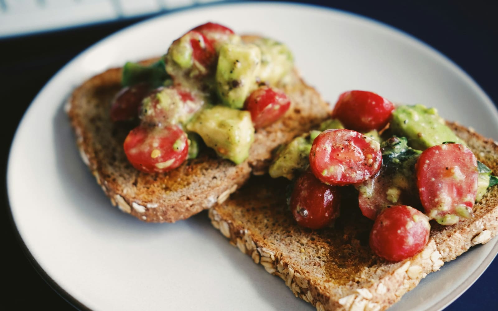 Two slices of seeded wholegrain toast topped with chopped avocado, halved cherry tomatoes and greens on a pale gray plate, close up
