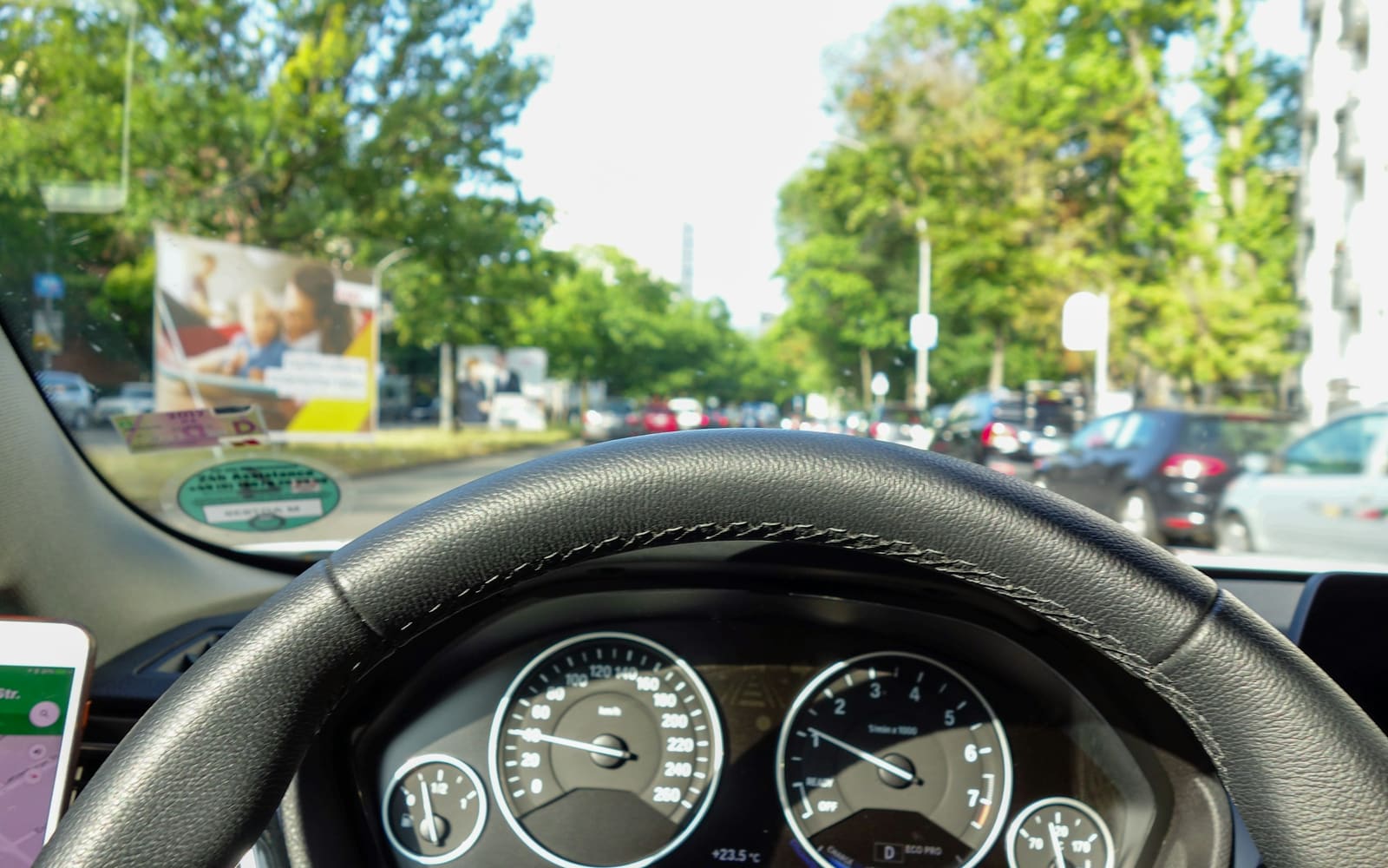 A driver’s view over a black leather steering wheel and dashboard dials toward a tree-lined city street with parked cars on a sunny day