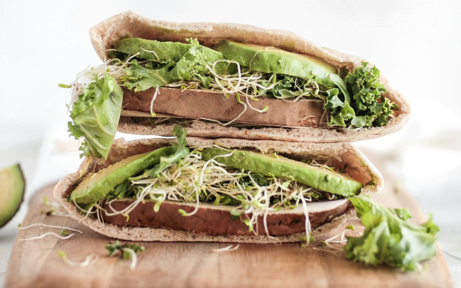 Two stuffed pita halves stacked on a wooden board, filled with sliced avocado, curly kale, alfalfa sprouts and a thick brown slice, against a white background