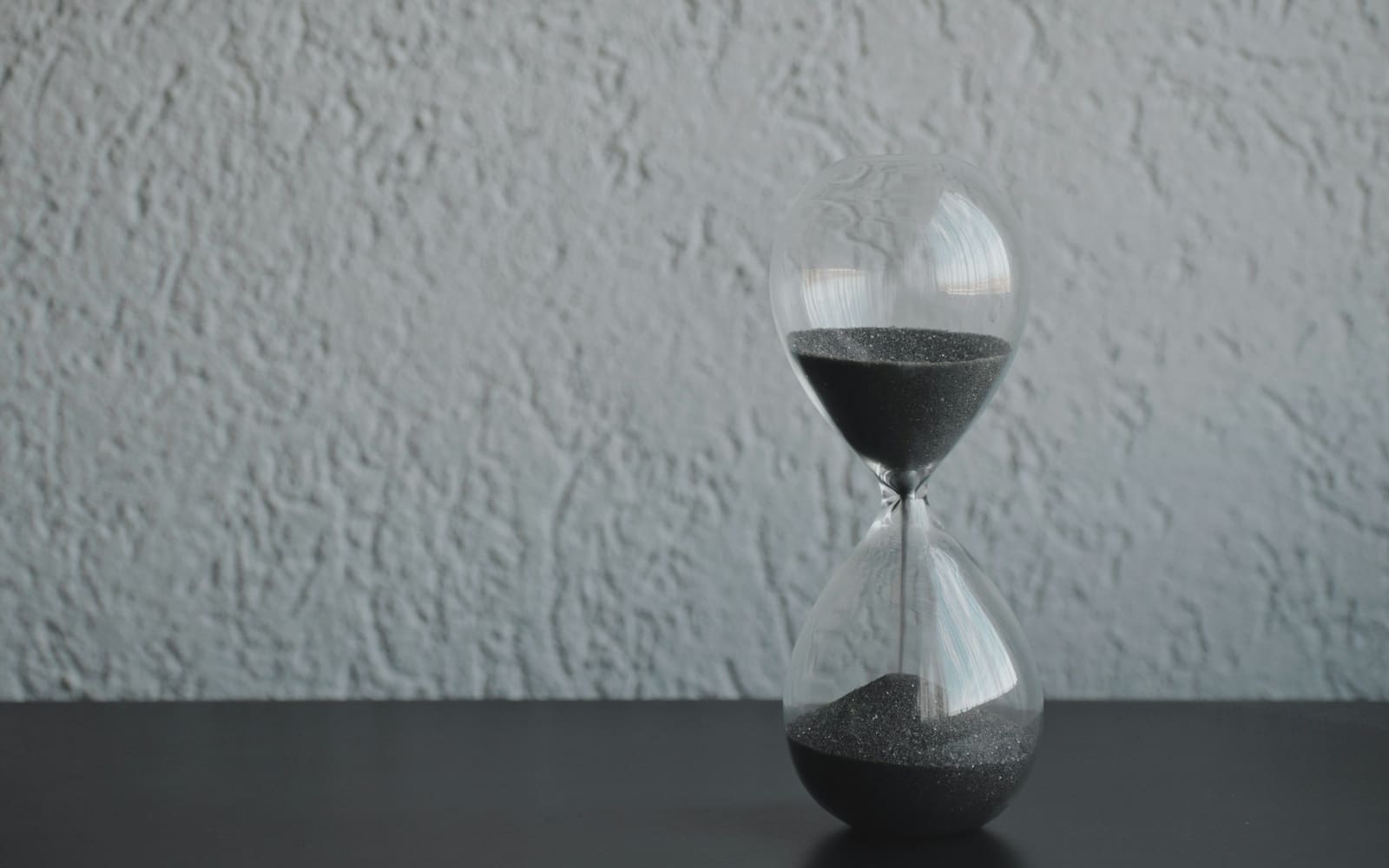 A glass hourglass with gray sand stands on a black table against a textured white wall, sand running from the top bulb into the bottom