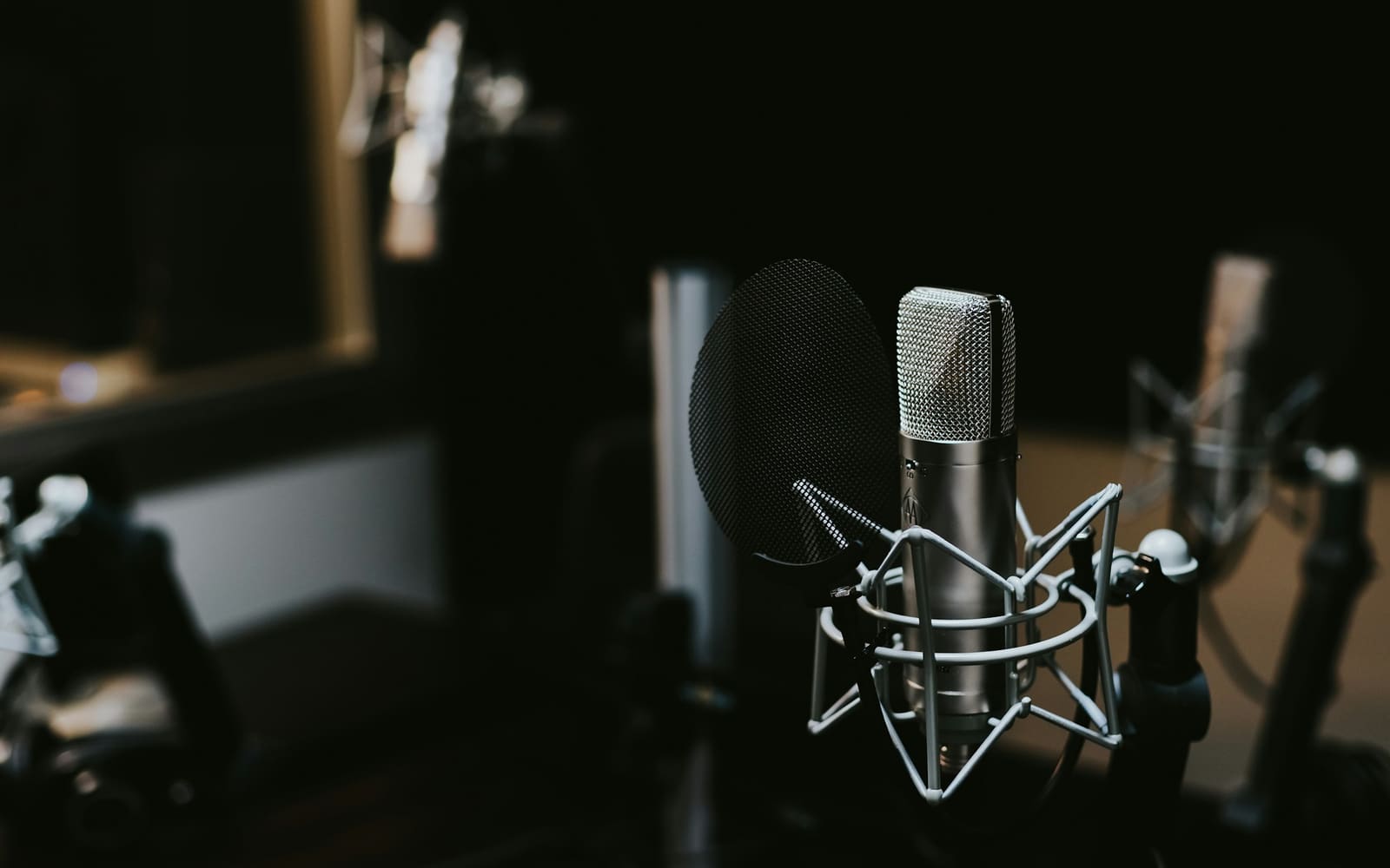 A silver studio microphone in a white shock mount beside a black mesh pop filter, in a dark recording room with more microphones blurred behind