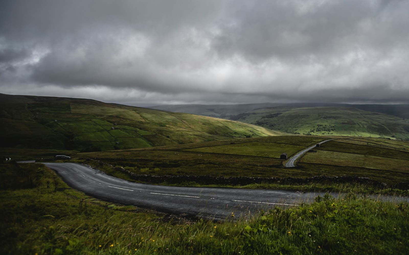 A wet country road curves across green moorland and stone-walled fields under heavy gray clouds, with a small barn and a car in the distance
