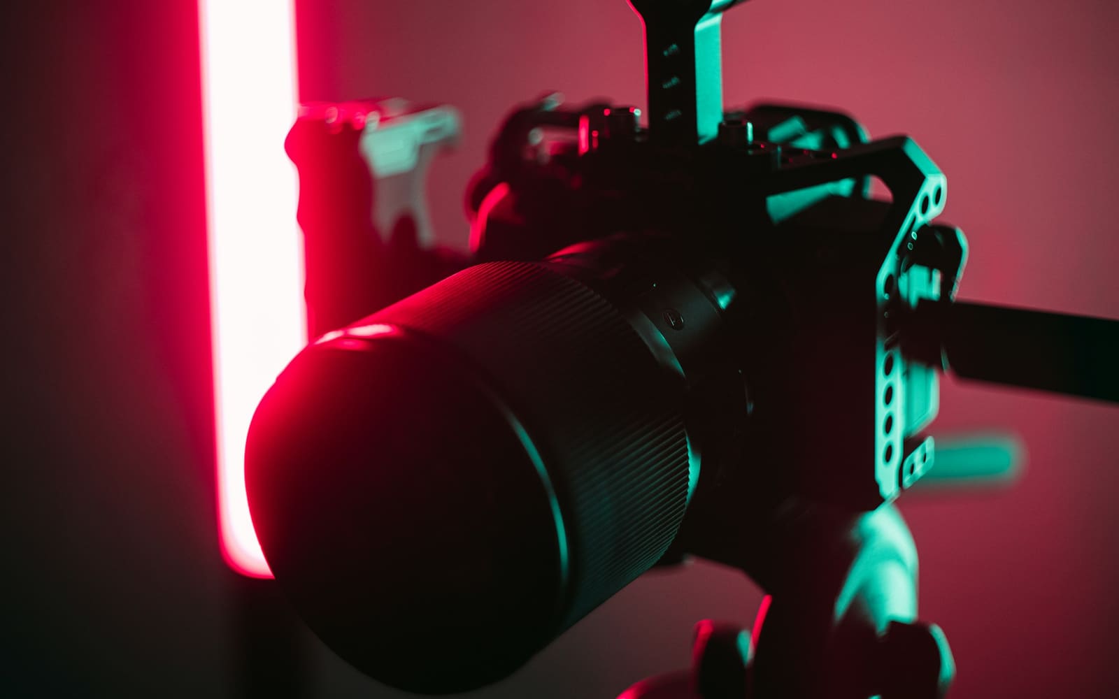 A video camera in a metal cage on a tripod, its large lens lit red and green by a glowing red light tube in a dark room