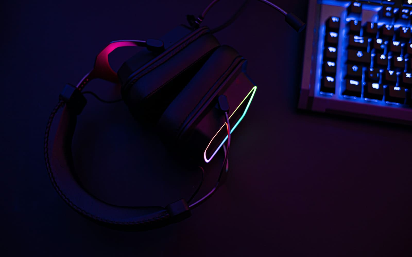 A black over-ear gaming headset with a glowing rainbow light strip lies on a dark desk beside a blue-backlit keyboard