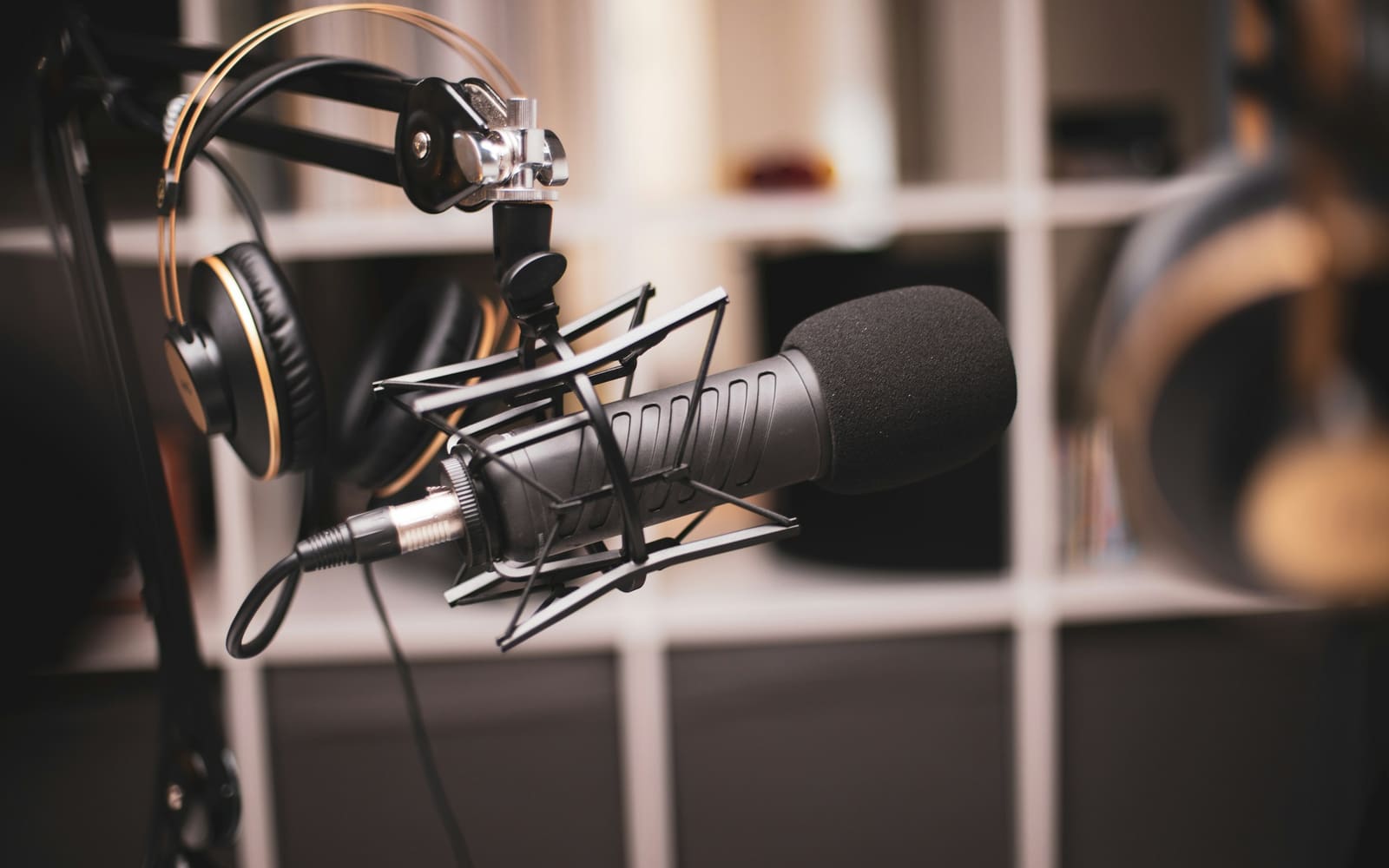 A black podcast microphone in a shock mount on a boom arm, with headphones hanging behind it in front of blurred white shelves