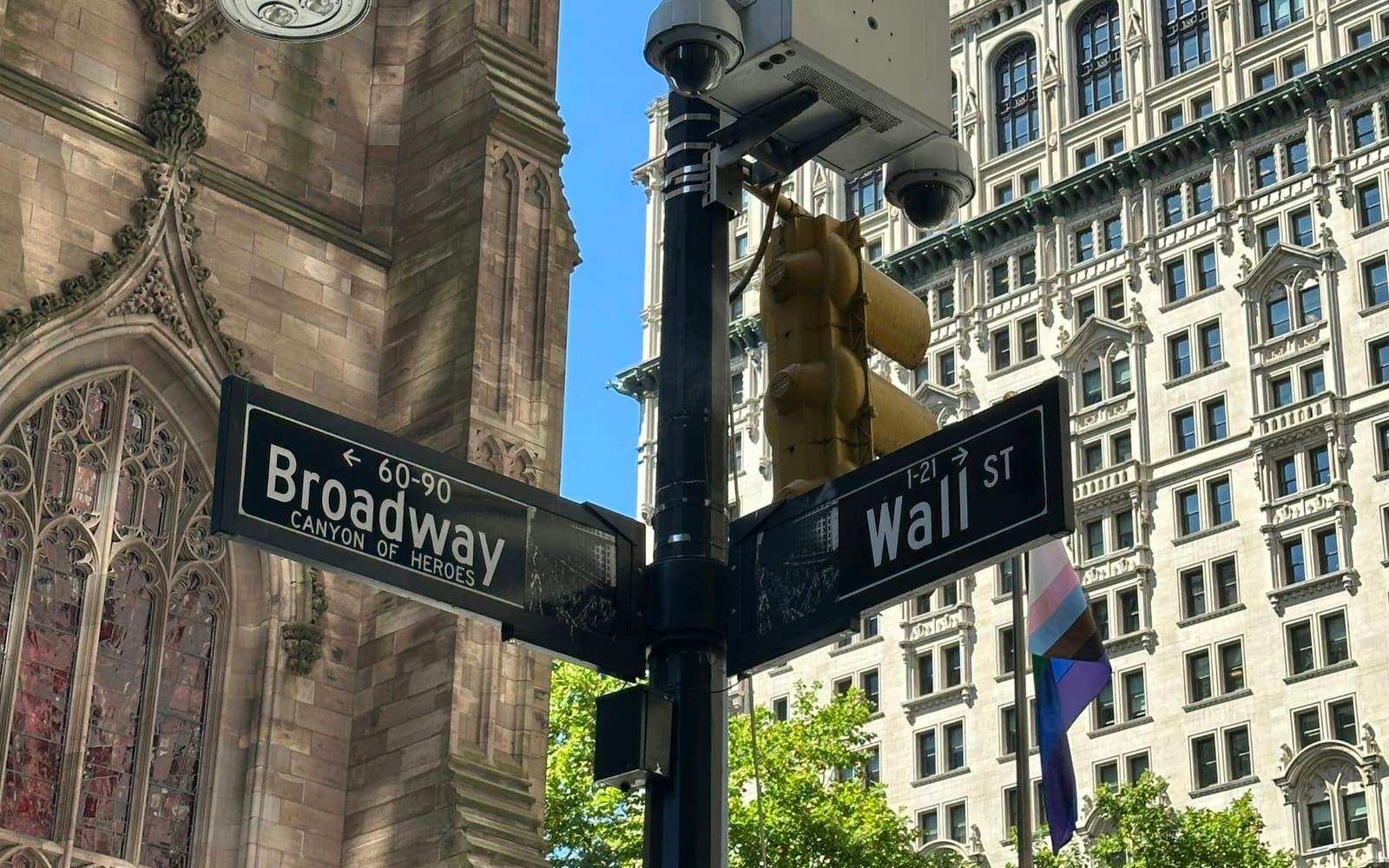 Black Broadway and Wall St street signs on a lamppost beside a Gothic church tower and an ornate office building under blue sky