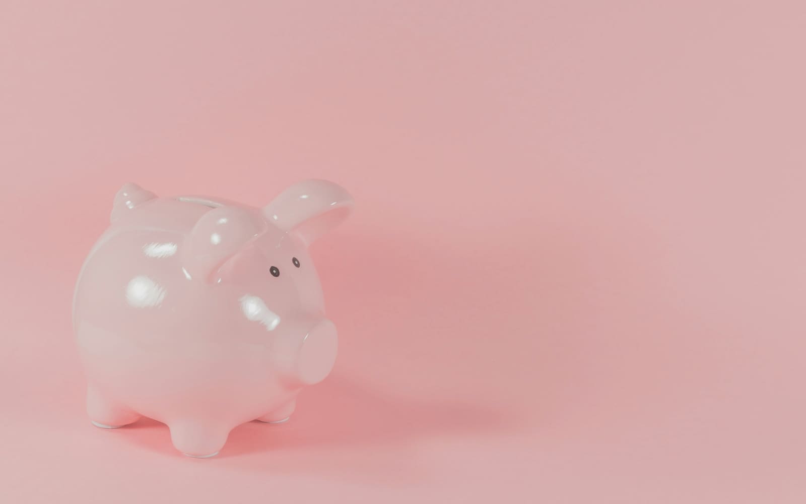 A glossy pale pink ceramic piggy bank stands on the left of a plain pink background, casting a soft shadow to the right