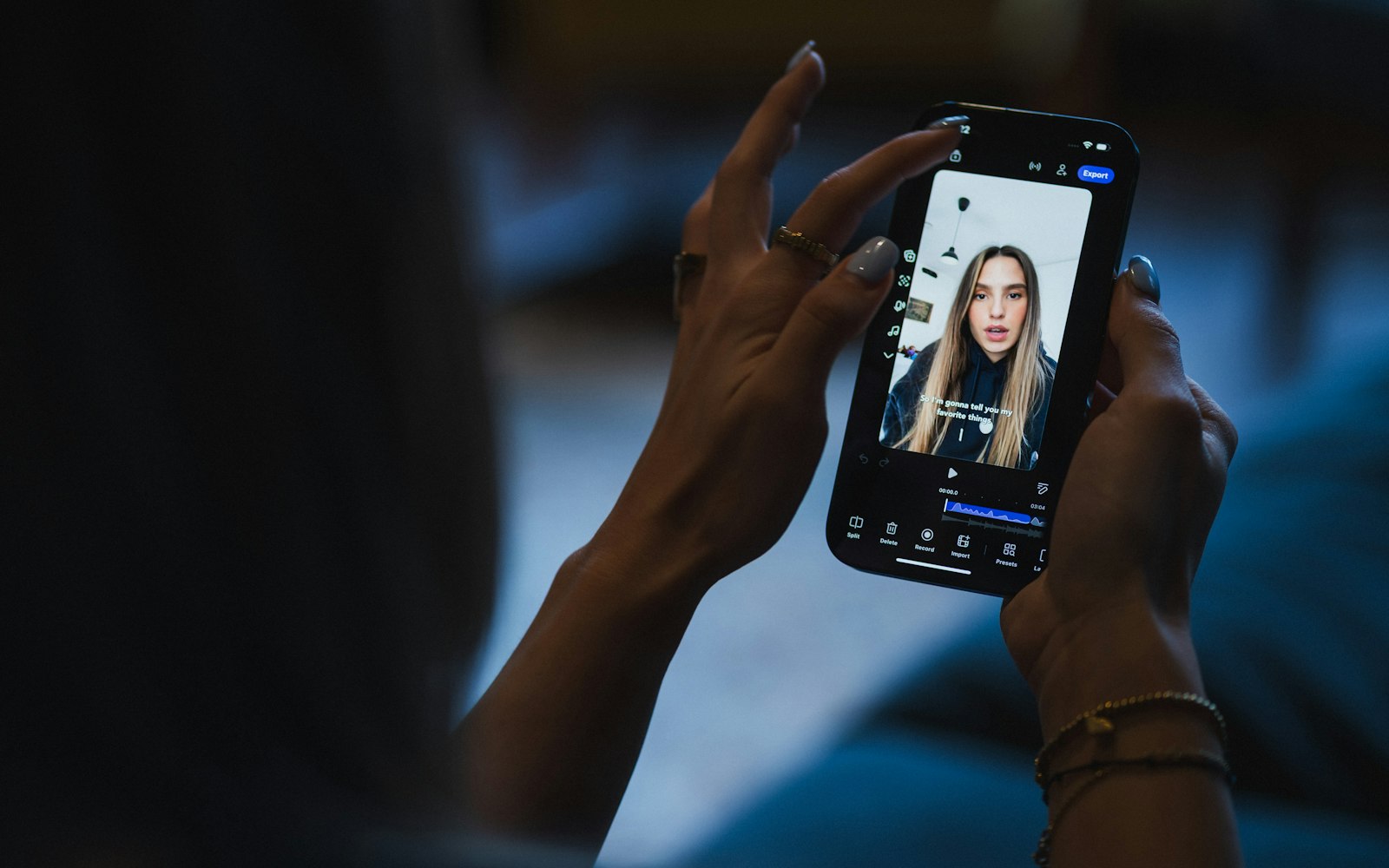 Hands with rings and painted nails hold a smartphone showing a vertical video of a young woman talking, with captions and an editing timeline below