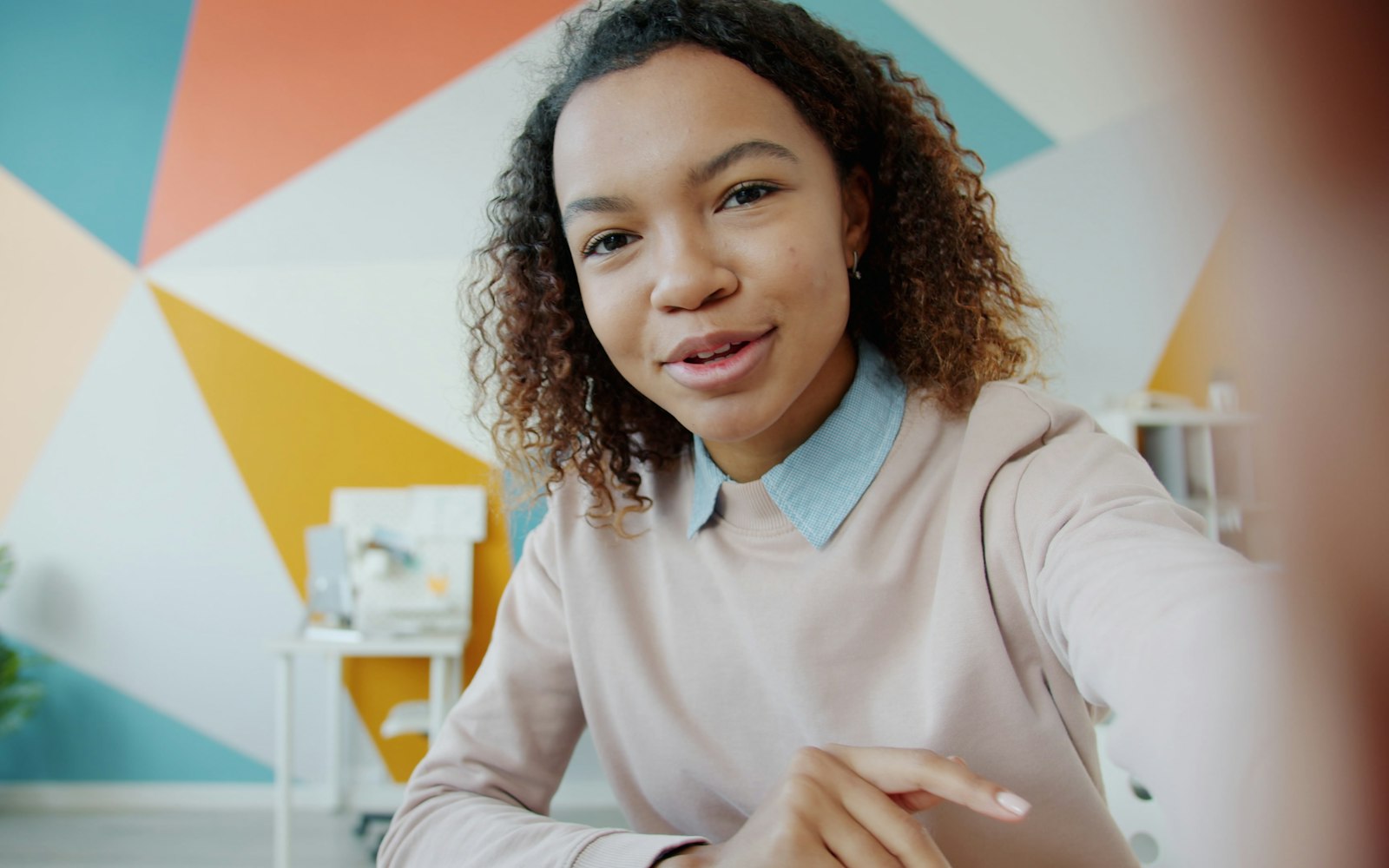 A young woman with curly hair, in a pink sweater over a collared shirt, talks into a camera held at arm’s length in front of a wall painted in colorful geometric shapes