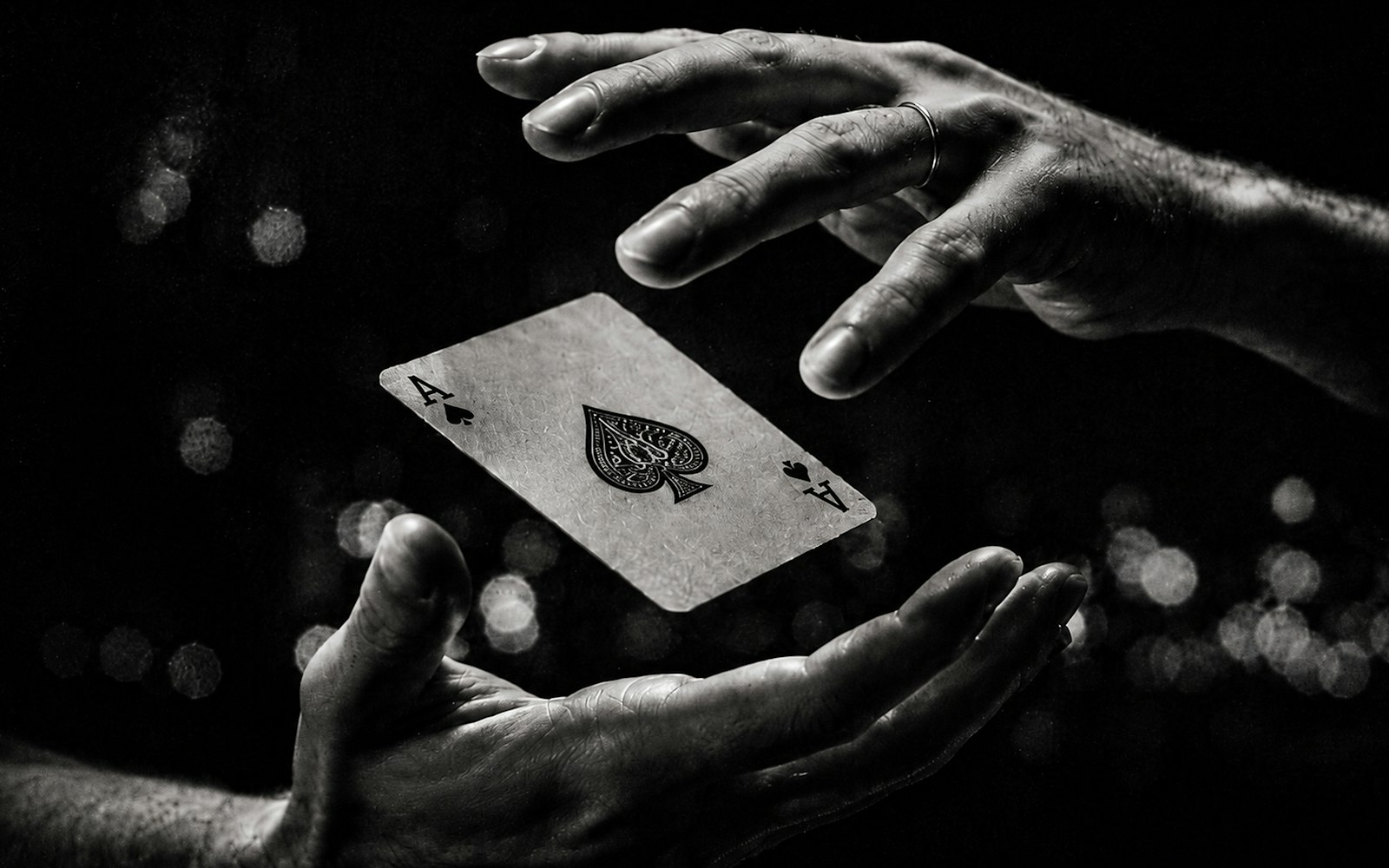 Black-and-white close-up of two hands, one with a ring, apparently levitating an ace of spades between them against a dark background with blurred lights
