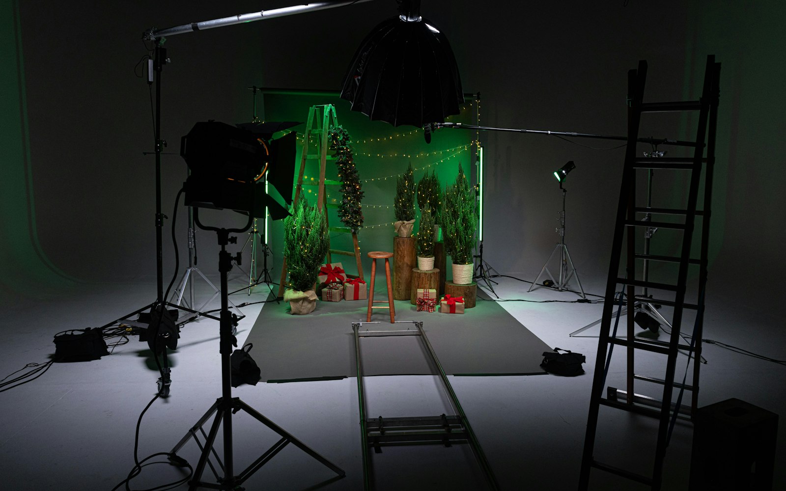 A dark film studio set with a green-lit backdrop, string lights, potted trees and wrapped gifts, surrounded by lights, ladders and a camera dolly track