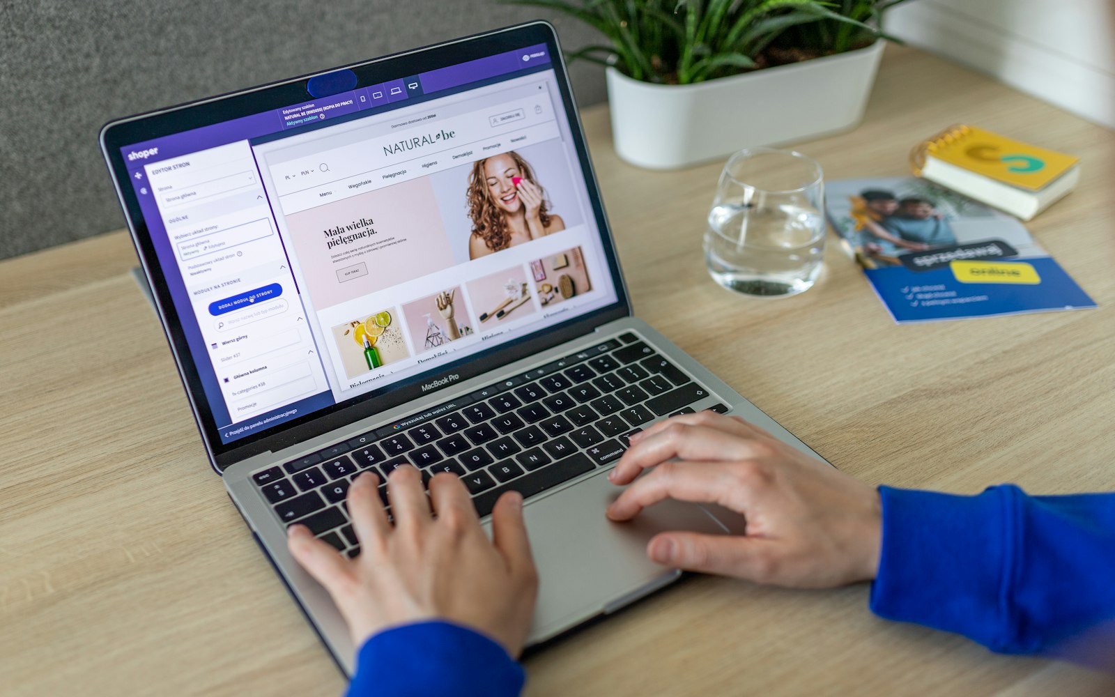 Hands typing on a laptop whose screen shows an online store page editor, with a skincare storefront preview beside the settings panel, a glass of water and a plant on the desk