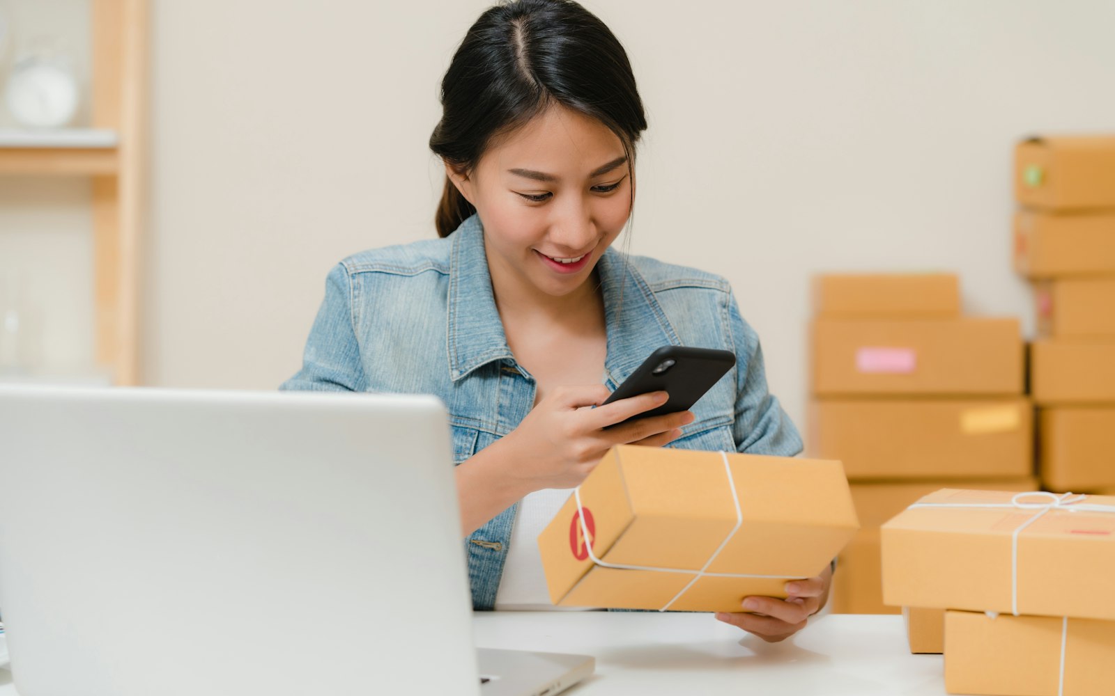 A woman in a denim jacket at a desk with an open laptop holds a small parcel tied with string and checks her phone, with stacked cardboard boxes behind her