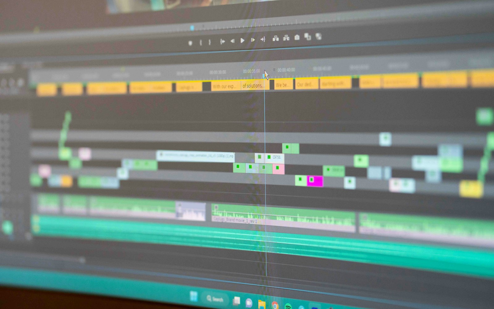 Close-up of a video editing timeline on a monitor, with a row of yellow caption clips along the top, colour-coded video clips below and green audio waveforms at the bottom