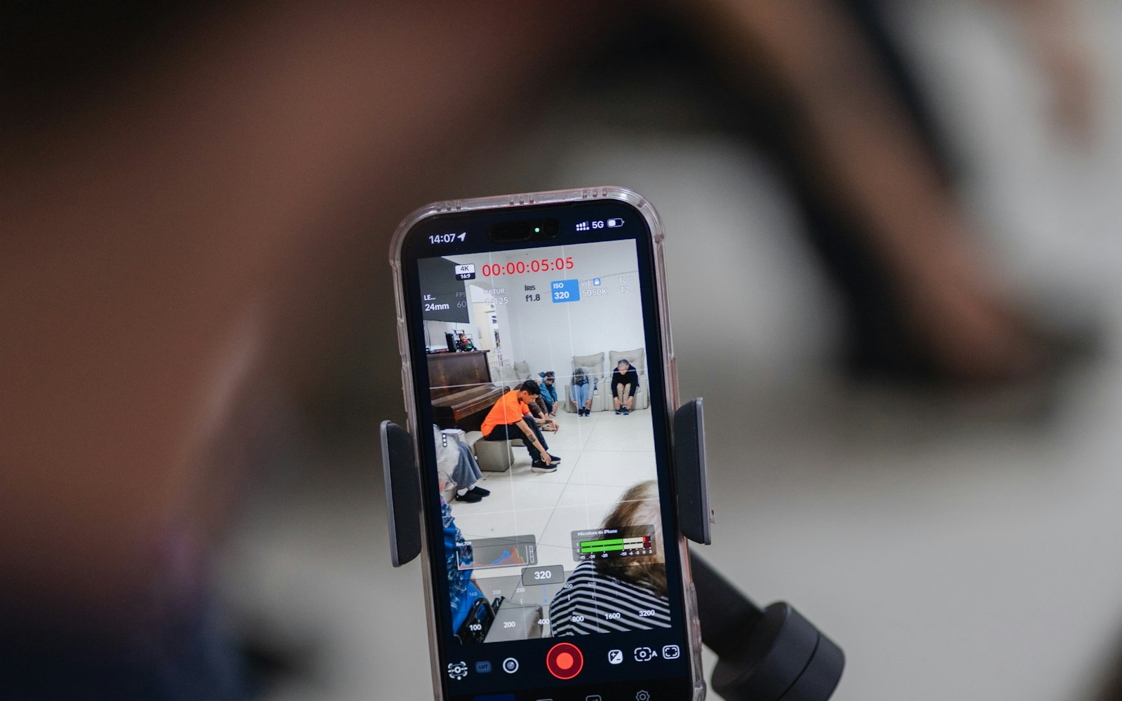 A smartphone in a gimbal clamp recording vertical video, its screen showing a five-second timecode and people seated along a wall, with the operator’s arm blurred in the foreground