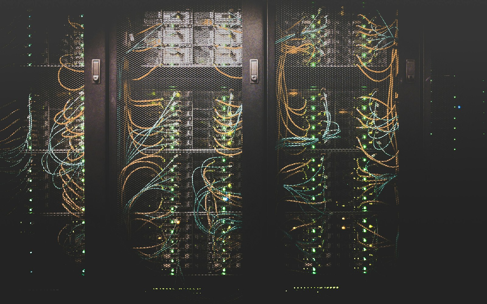 Three server cabinets with mesh doors in a dark data centre, orange and blue cables looping between rows of green status lights
