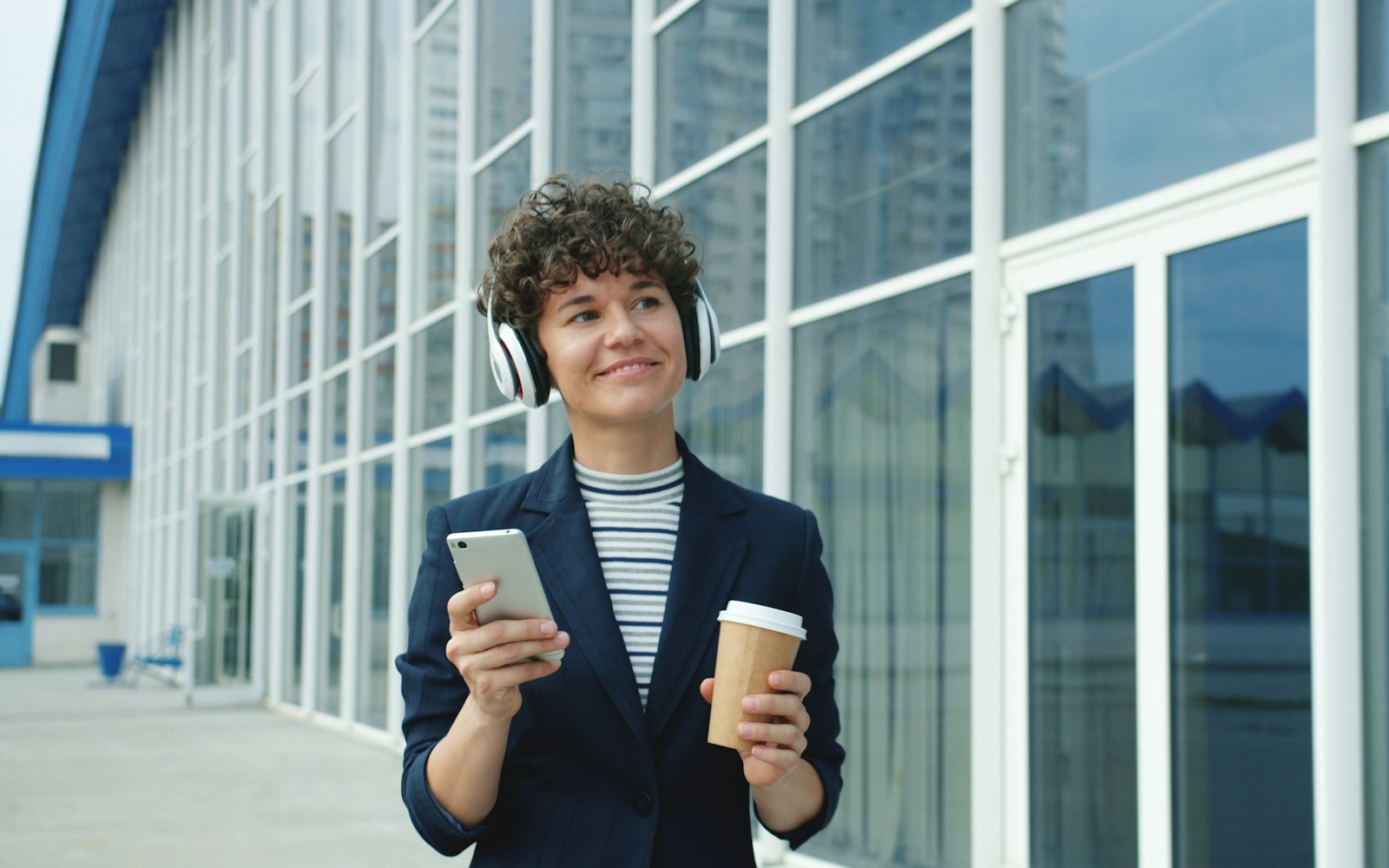 A smiling woman in a navy blazer and white headphones walking past a glass office building, holding a phone and a takeaway coffee