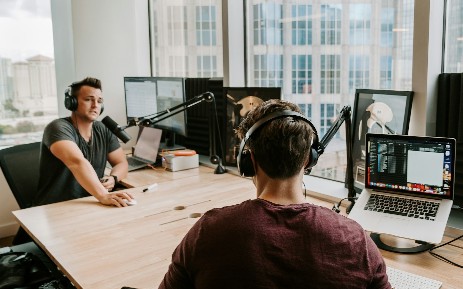 Two men in headphones recording a podcast across a wooden desk, each with a microphone on a boom arm, with a laptop and city windows behind them