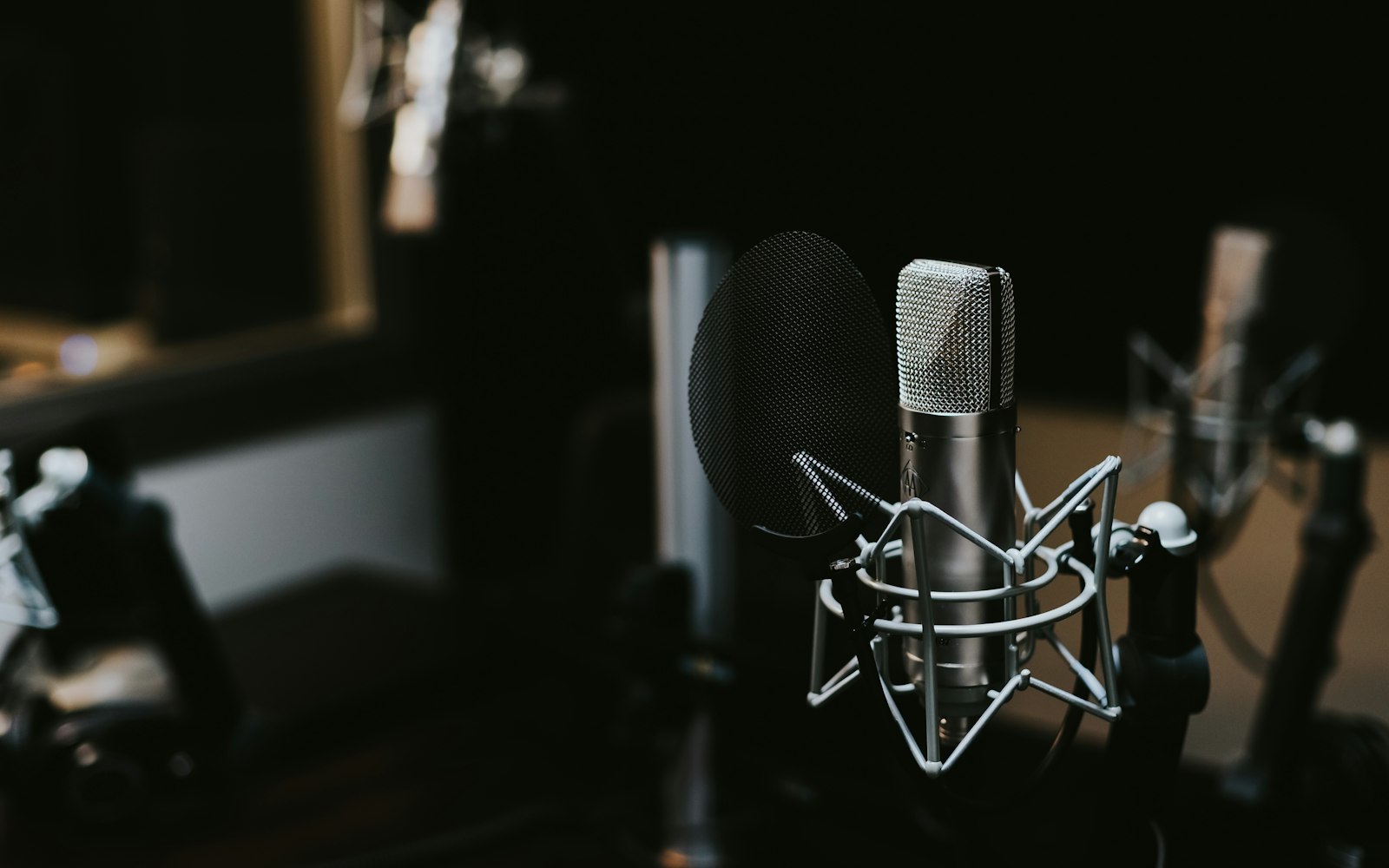 A silver condenser microphone in a shock mount with a mesh pop filter, in a dark recording studio with other microphones blurred behind it