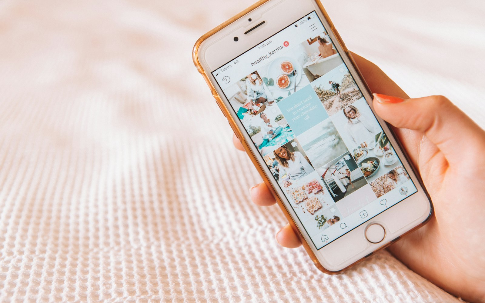 A hand holds a gold-cased iPhone showing an Instagram profile grid of pastel food, beach and exercise photos on a white blanket