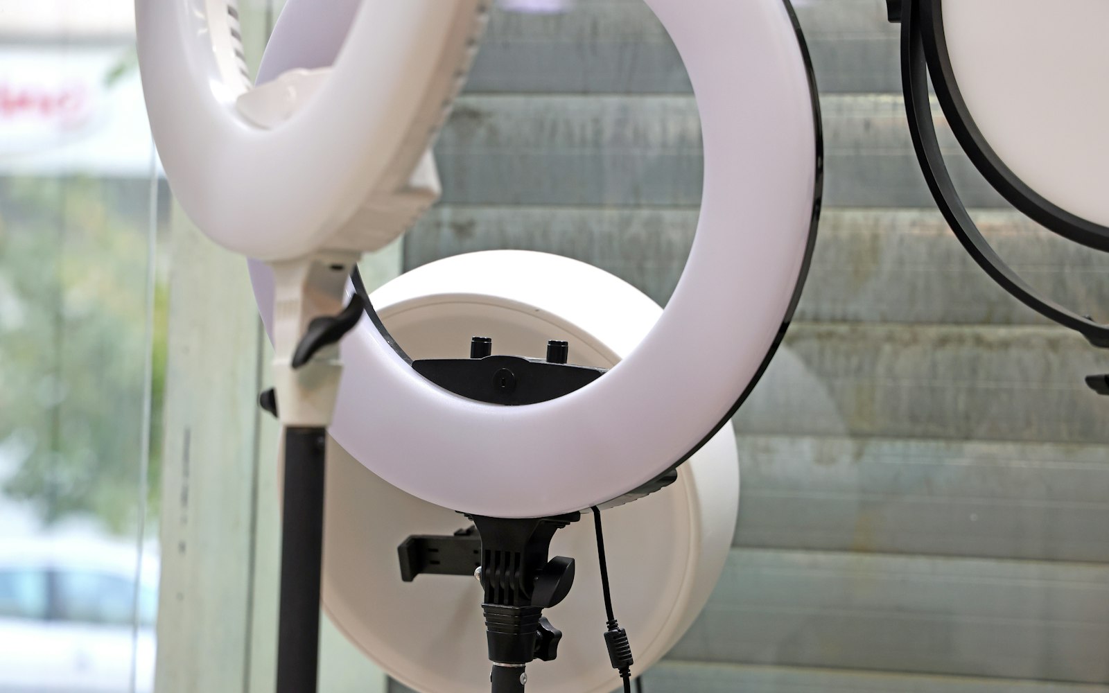 Three white LED ring lights on black stands beside a window with half-open blinds, the kind of setup used to film to camera