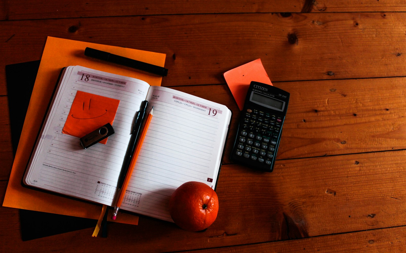 Overhead view of an open diary on a wooden table, with a pencil and a pen laid across the page, an orange sticky note with a smiley face, a USB stick, a scientific calculator and a tangerine