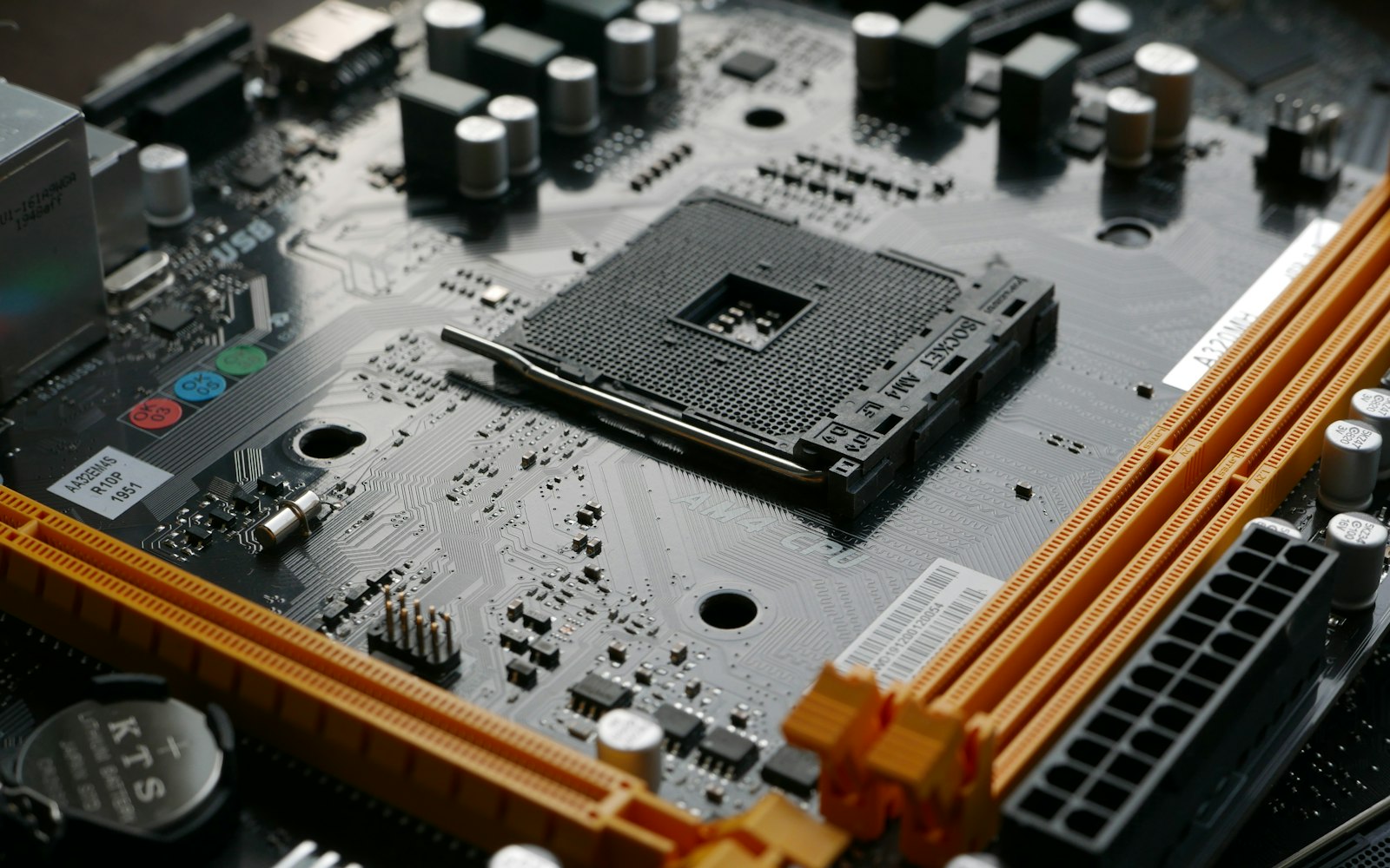 Close-up of a bare computer motherboard with an empty CPU socket, orange memory slots, a coin-cell battery and rows of capacitors