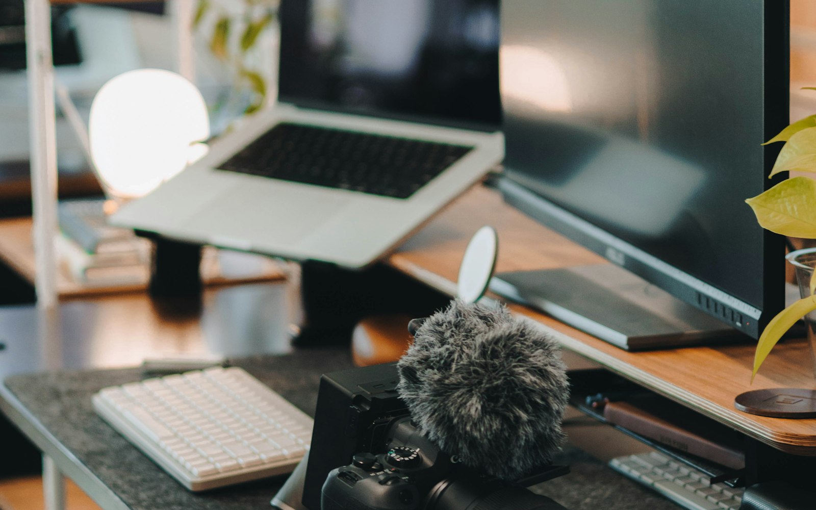 A desk with a laptop on a stand, a large monitor, a compact white keyboard and a camera fitted with a furry microphone windscreen in the foreground