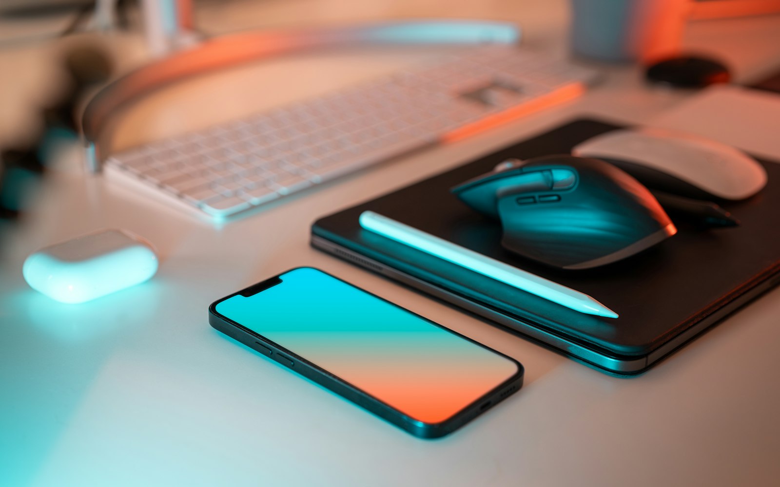 A smartphone, a tablet with a stylus, a wireless mouse, an earbuds case and a keyboard laid out on a white desk under teal and orange light