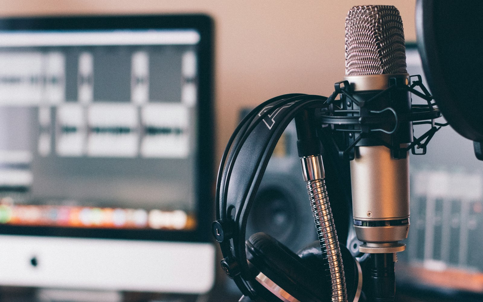 A condenser microphone in a shock mount with headphones hanging from its stand, in front of a blurred monitor showing a video-editing timeline